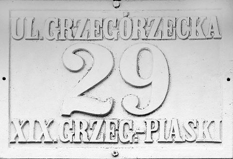 Fot. 27. W roku 1881 Rada Miasta Krakowa, utrzymując dotychczasowy podział na 8 dzielnic katastralnych (jedynie Dzielnicę I w miejsce nazwy „Kraków” nazwano „Śródmieściem”), wprowadziła nowe zasady ponumerowania orientacyjnego domów w mieście. Zasady te przetrwały do dzisiaj: numeracja domów ulicami (a nie dzielnicami katastralnymi, jak poprzednio), początek numerowania od strony bliższej Rynkowi Głównemu (na Stradomiu bliższej ul. Stradomskiej, na Kazimierzu bliższej ul. Krakowskiej), numery nieparzyste po prawej stronie. Wprowadzono jednolite dla całego miasta kolory tabliczek z nazwami ulic i tabliczek orientacyjnych z numerami domów: litery i cyfry koloru czarnego, tło białe, obwódki tablic niebieskie (DzRMK 1881, nr 1, s. 2–3). Przydzielone wówczas nowe numery domów pozostały już z reguły niezmienione do naszych czasów (zmiany w przynależności dzielnicowej ulic nie miały znaczenia dla numeru domu), natomiast sam wzór tabliczek wprawdzie był aktualny jeszcze w okresie okupacji, jednak już w okresie międzywojennym podlegał wymianie na znany nam co do kolorystyki z codzienności wzór nowy, z białymi literami i cyframi na niebieskim tle, z białą obwódką. Powyżej: 4) tabliczka już z okresu Wielkiego Miasta Krakowa (Grzegórzki–Piaski przyłączono do Krakowa w 1910 r., ustanawiając dla tej dawnej gminy wiejskiej odrębną dzielnicę katastralną miasta o numerze XIX).