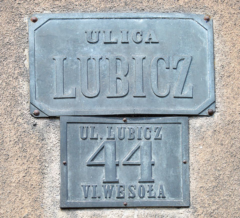 Fot. 28. W roku 1881 Rada Miasta Krakowa, utrzymując dotychczasowy podział na 8 dzielnic katastralnych (jedynie Dzielnicę I w miejsce nazwy „Kraków” nazwano „Śródmieściem”), wprowadziła nowe zasady ponumerowania orientacyjnego domów w mieście. Zasady te przetrwały do dzisiaj: numeracja domów ulicami (a nie dzielnicami katastralnymi, jak poprzednio), początek numerowania od strony bliższej Rynkowi Głównemu (na Stradomiu bliższej ul. Stradomskiej, na Kazimierzu bliższej ul. Krakowskiej), numery nieparzyste po prawej stronie. Wprowadzono jednolite dla całego miasta kolory tabliczek z nazwami ulic i tabliczek orientacyjnych z numerami domów: litery i cyfry koloru czarnego, tło białe, obwódki tablic niebieskie (DzRMK 1881, nr 1, s. 2–3). Przydzielone wówczas nowe numery domów pozostały już z reguły niezmienione do naszych czasów (zmiany w przynależności dzielnicowej ulic nie miały znaczenia dla numeru domu), natomiast sam wzór tabliczek wprawdzie był aktualny jeszcze w okresie okupacji, jednak już w okresie międzywojennym podlegał wymianie na znany nam co do kolorystyki z codzienności wzór nowy, z białymi literami i cyframi na niebieskim tle, z białą obwódką. Powyżej: 2) zachowana i służąca do dzisiaj para tabliczek: z nazwą ulicy i z numerem domu.